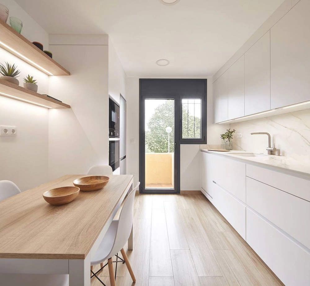 Cuina moderna i funcional amb mobiliari blanc, taula de fusta i il·luminació natural a Osona
